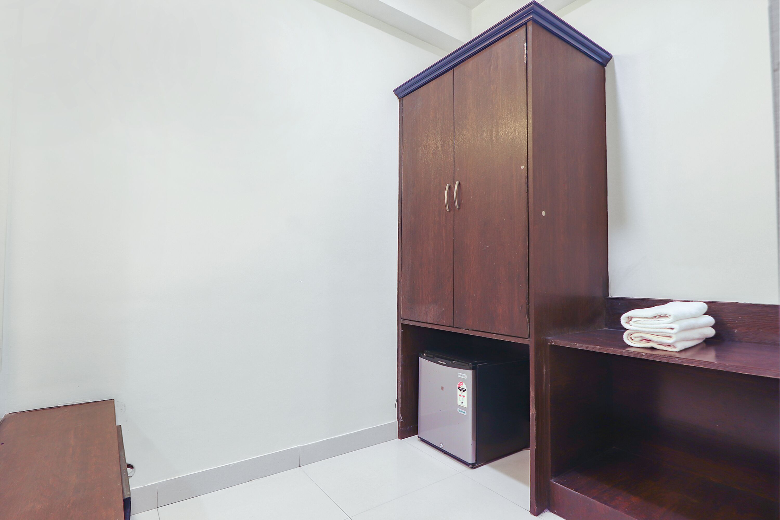 A small, dark wooden wardrobe with a built-in mini-fridge and a shelf with folded towels.