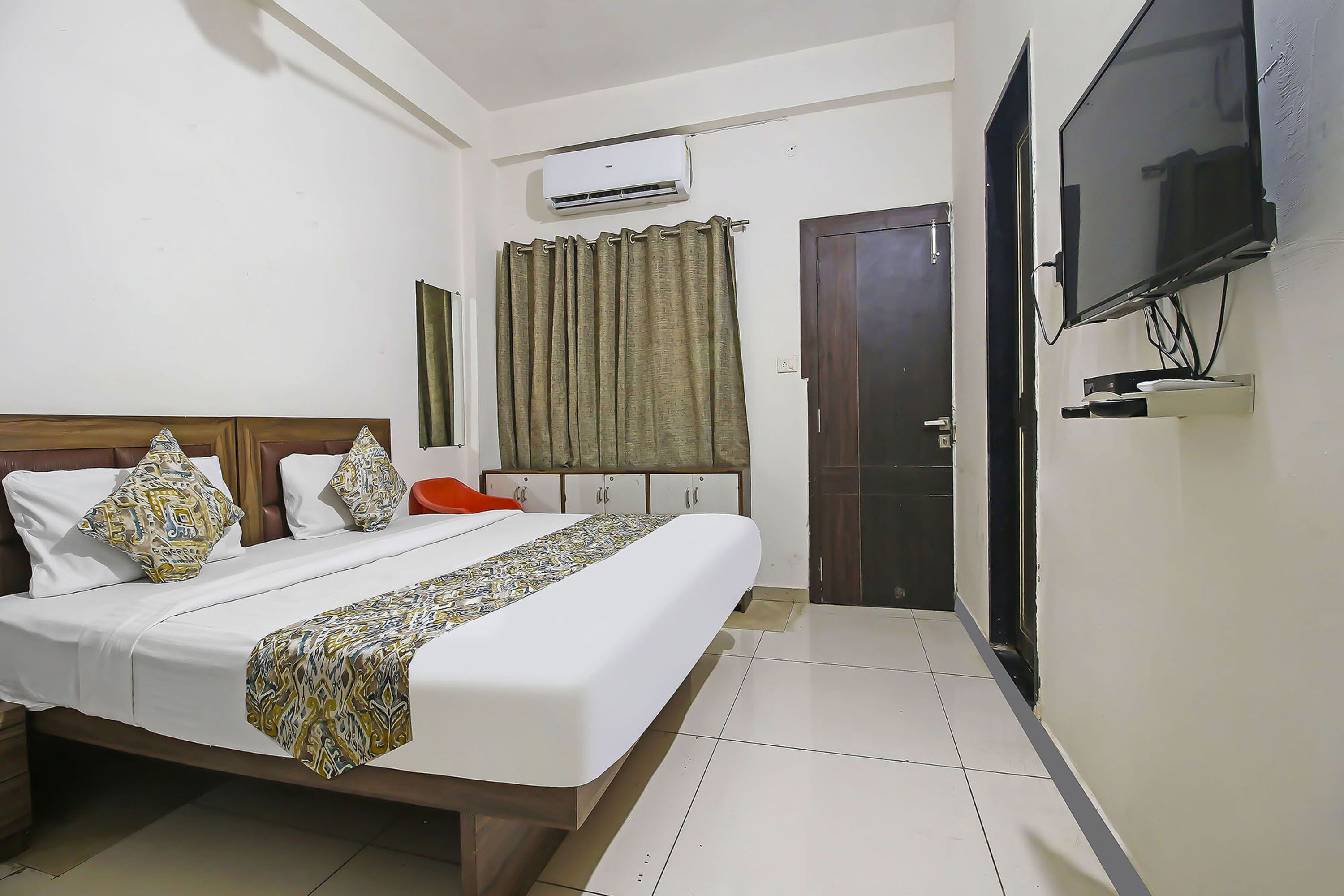 A hotel room with a bed, an air conditioner, a TV, a door, and a curtain.