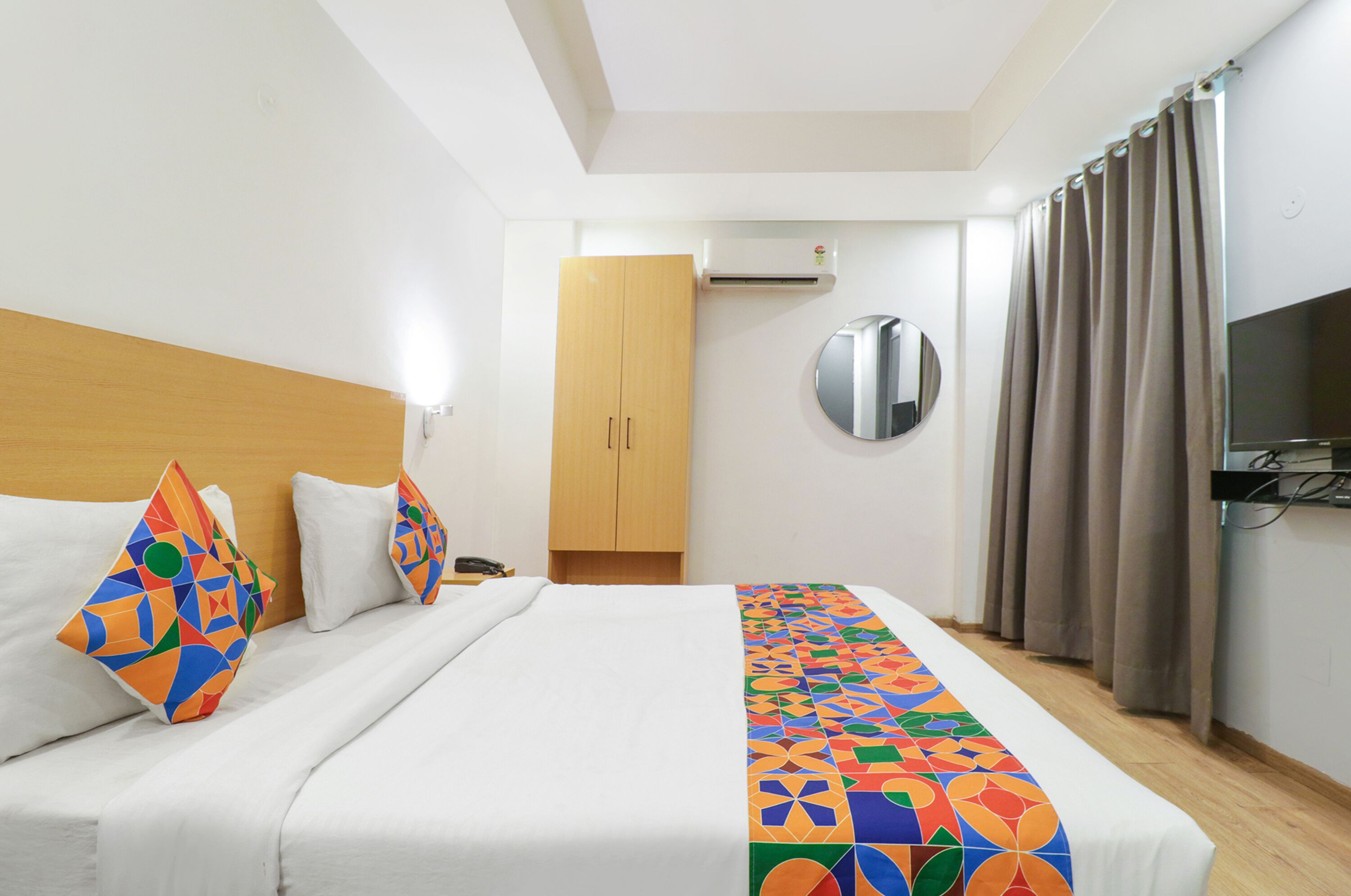 A hotel room with a bed, a wardrobe, an air conditioning unit, a round mirror, and a television.