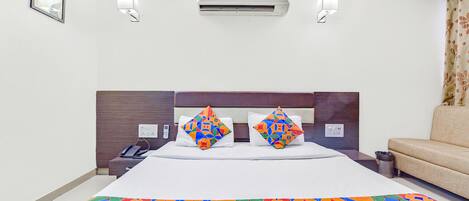 Quarto de hotel com uma cama, duas almofadas, mesa de cabeceira com telefone, ar condicionado, lixeira e um sofá.