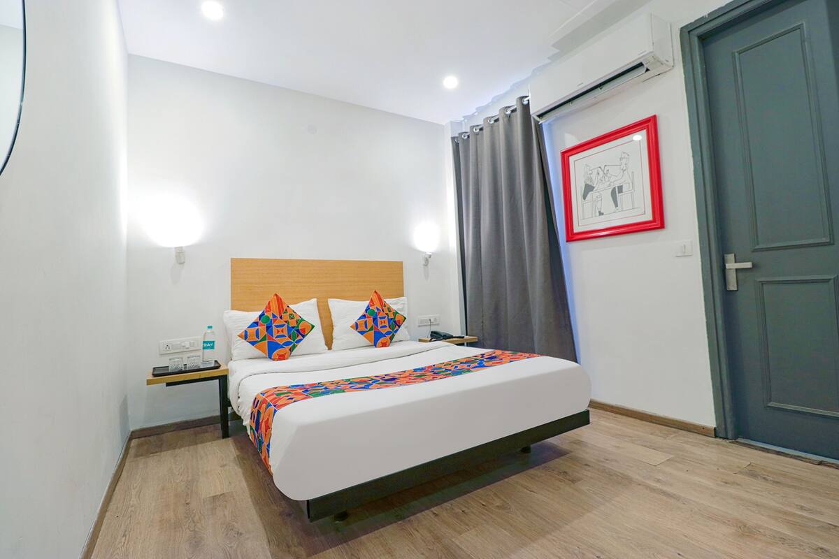 A neatly made bed with a patterned bedspread, a bedside table with a bottle and a phone, a framed picture on the wall, and a door to the right.