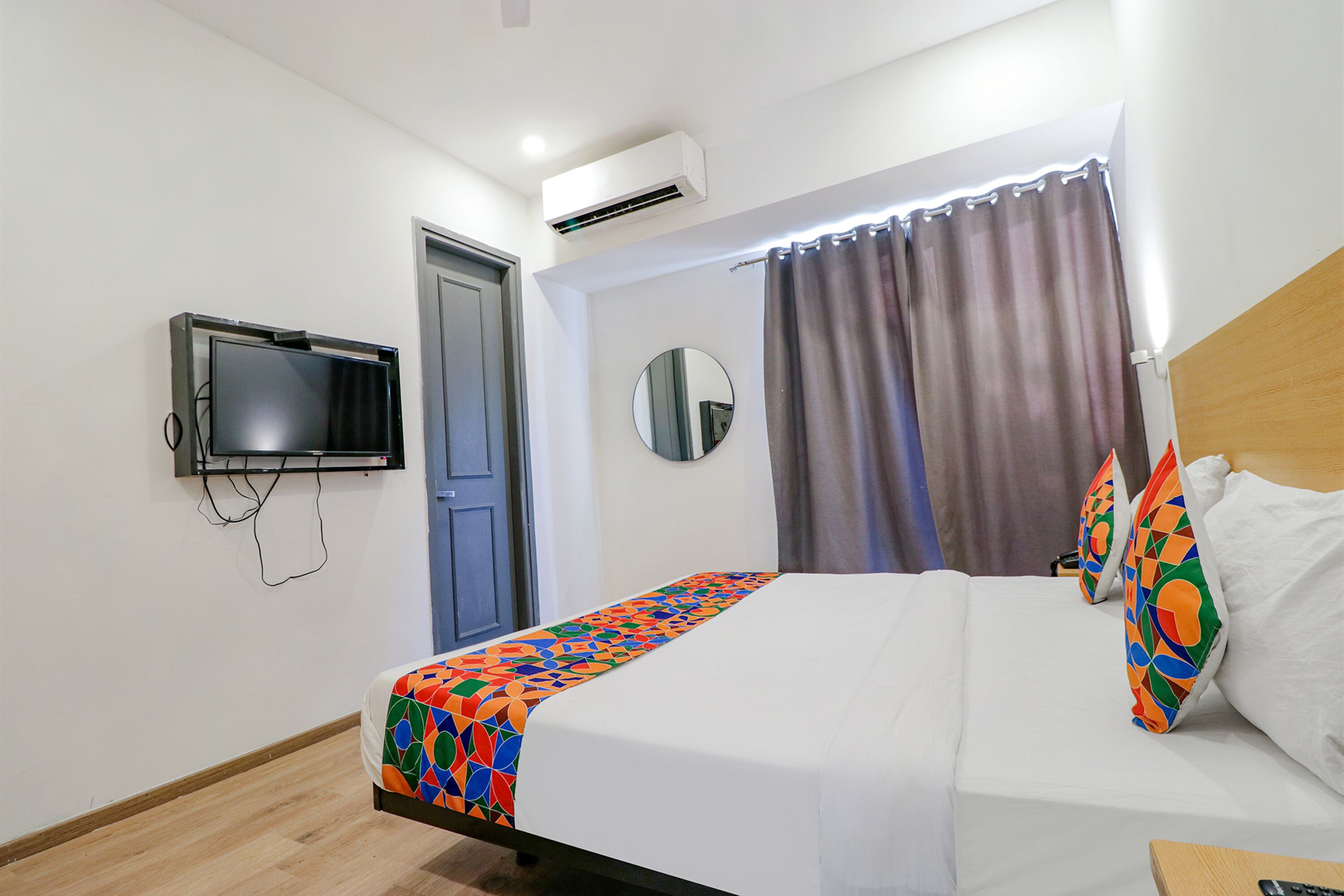 Ein Hotelzimmer mit einem Bett, einem an der Wand befestigten Fernseher, einer Klimaanlage, einer Tür und einem Fenster mit Vorhängen.