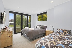 A modern bedroom with a large bed, a second bed, a wooden nightstand, a desk, and a large window with a view of greenery.