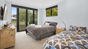 Quarto moderno com uma cama grande, uma cama menor, uma mesinha de madeira e uma janela ampla com vista para o verde.
