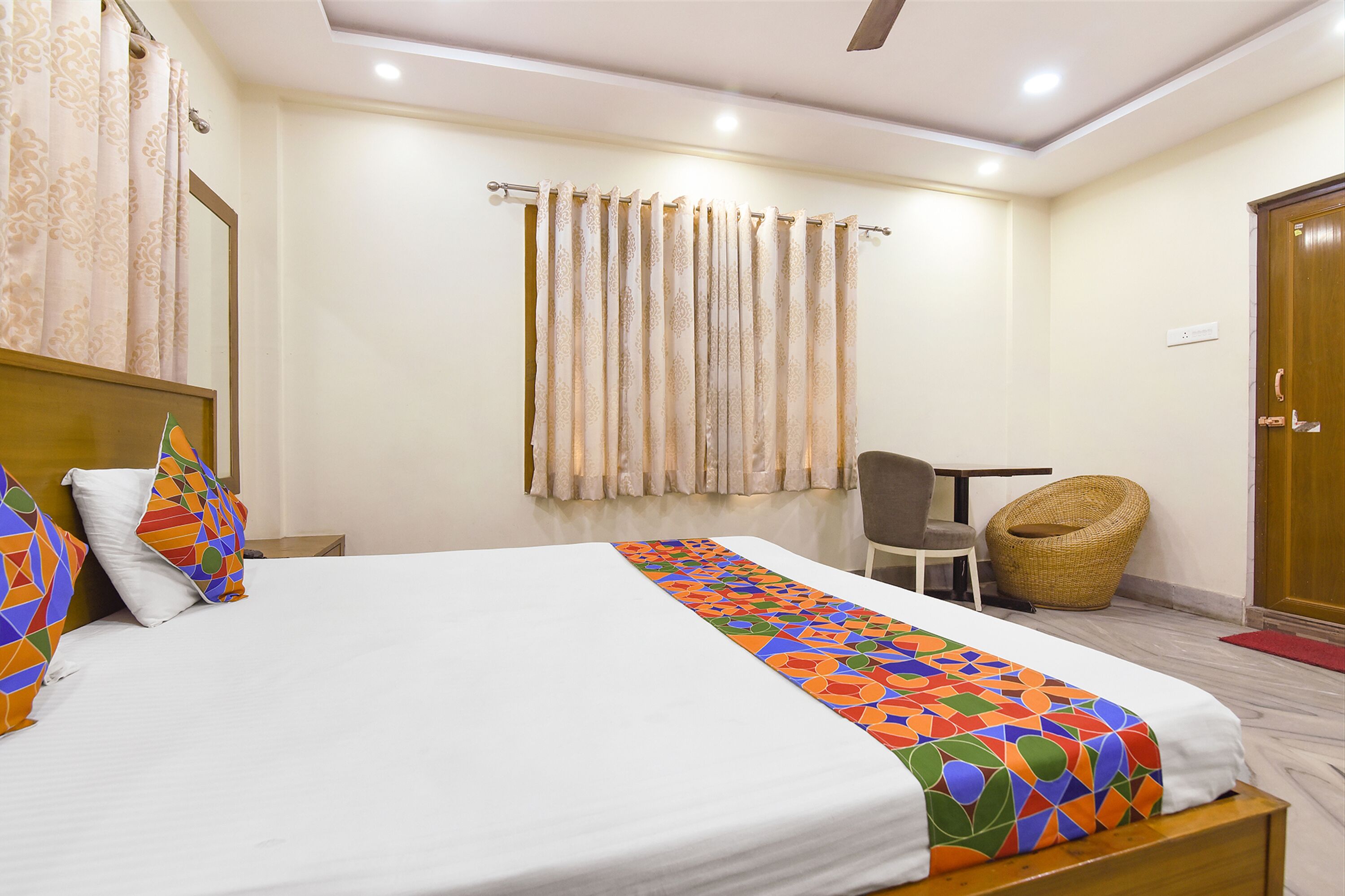 A hotel room with a bed, a chair, a wicker chair, a wooden door, and a curtain.