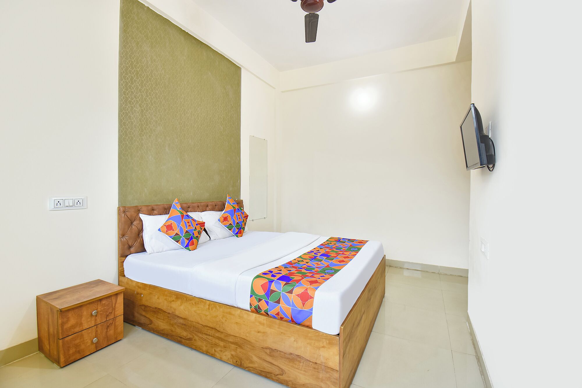 A neatly made bed with a wooden frame and colorful bedding, a small wooden nightstand, a ceiling fan, and a wall-mounted TV.
