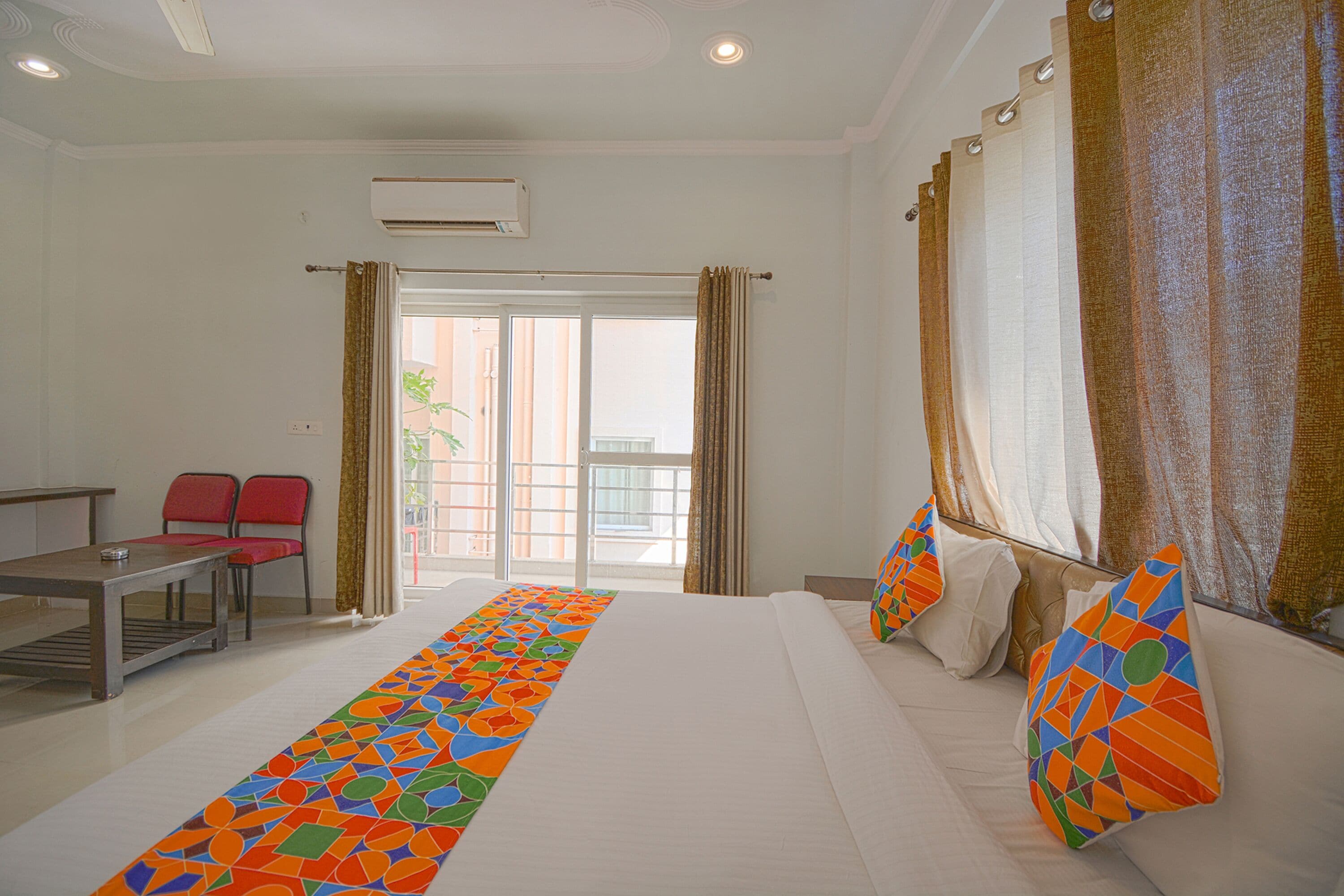 Habitación de hotel con cama, escritorio, dos sillas, ventana con cortinas y un corredor colorido.