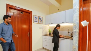 A modern kitchen with white cabinets, a countertop, and a sink. There is a door with a sign above it and a small potted plant on the counter. - Fabhotel Kings Suites (Bengaluru)