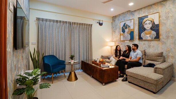 A living room with a blue chair, a brown sofa, a coffee table, and a ceiling fan. There are two paintings on the wall and a TV mounted on the wall. - Fabhotel Kings Suites (Bengaluru)