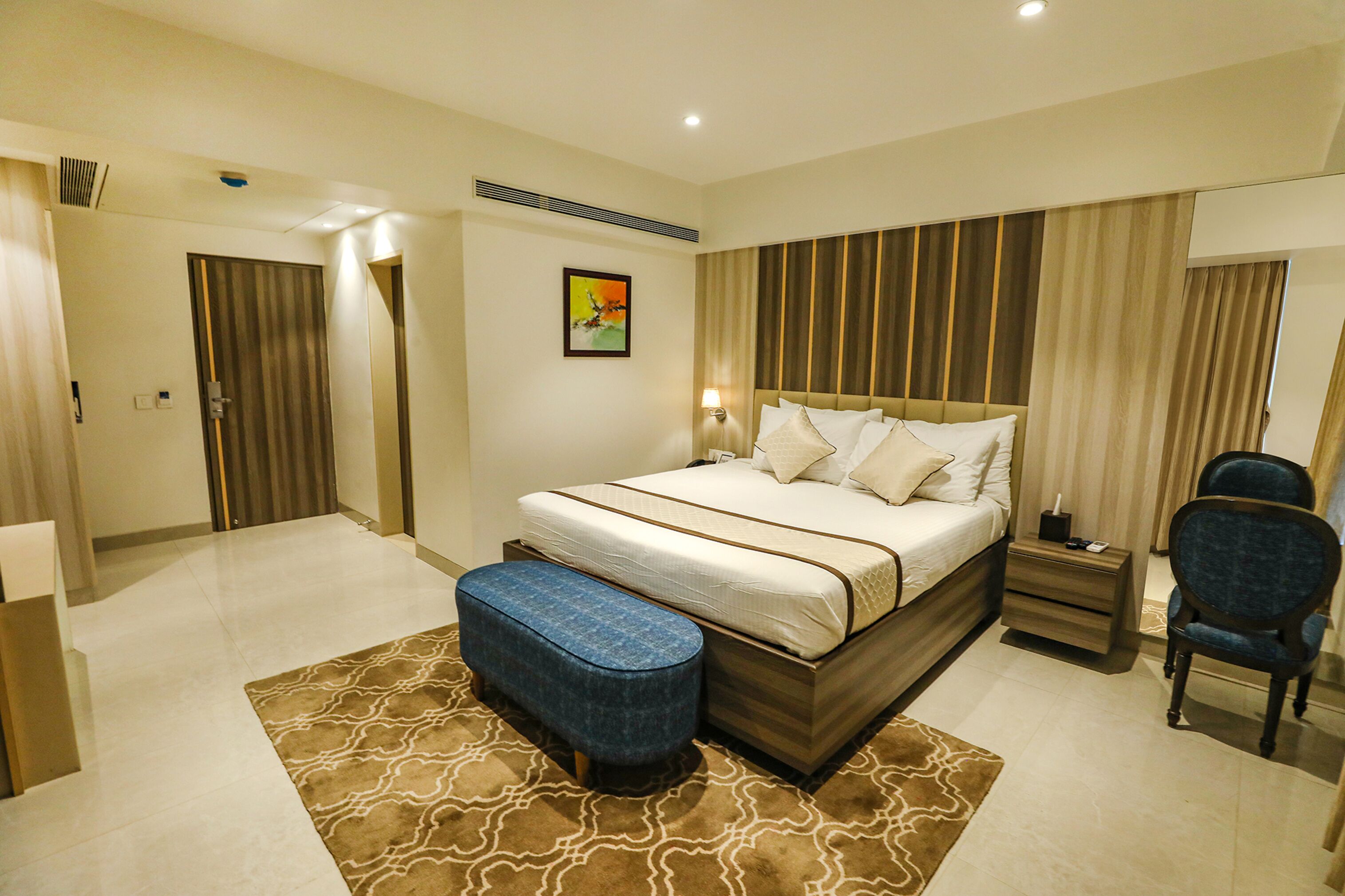 A hotel room with a large bed, a desk with a chair, a small ottoman, and a picture on the wall.