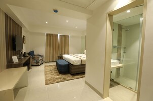 Una habitación de hotel moderna con cama, escritorio, sofá, ducha con mampara de vidrio y televisor de pantalla plana.