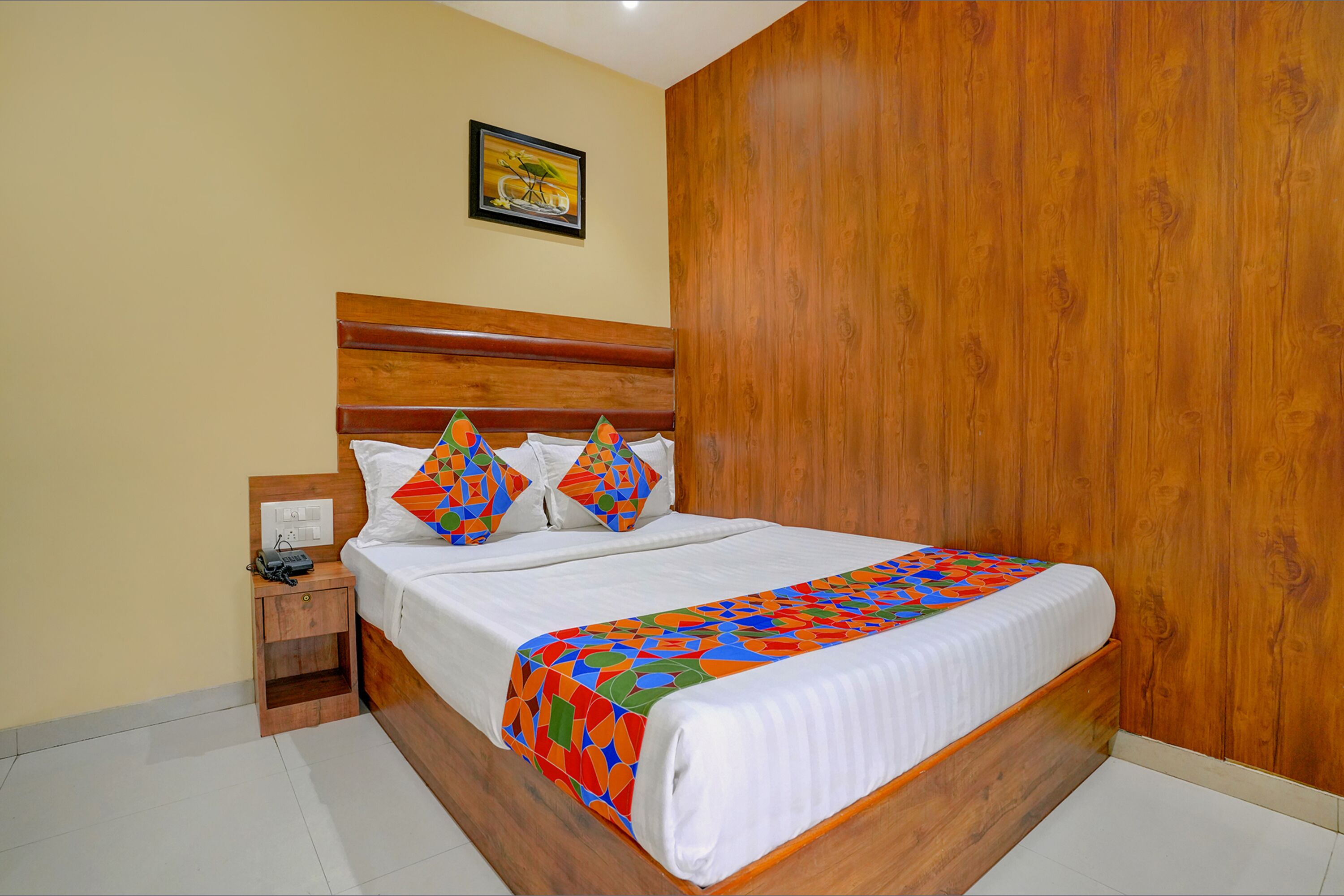 A neatly made bed with a colorful geometric-patterned bedspread, a wooden headboard, and a small bedside table with a phone.
