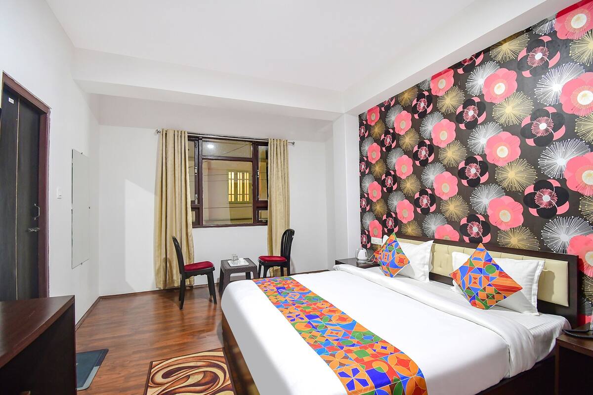 A hotel room with a bed, a desk, a chair, a window with curtains, and a wall with a floral pattern.