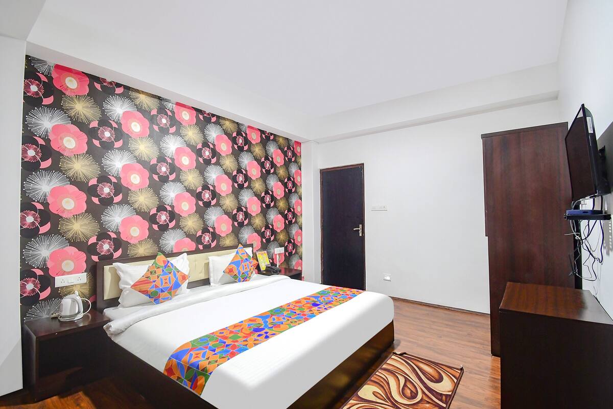 A hotel room with a large bed, a colorful bedspread, a dark wooden wardrobe, and a wall with a floral pattern.