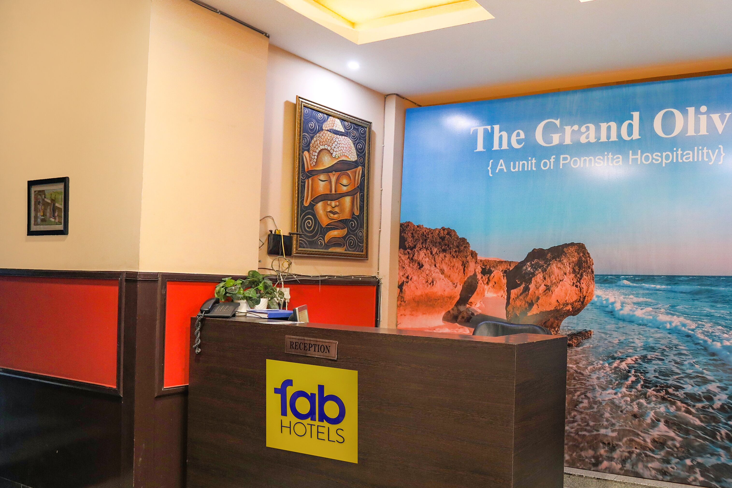 A hotel reception desk with a "Reception" sign, a "Tab Hotels" logo, a Buddha painting, and a large poster of "The Grand Olivia" with a coastal view.