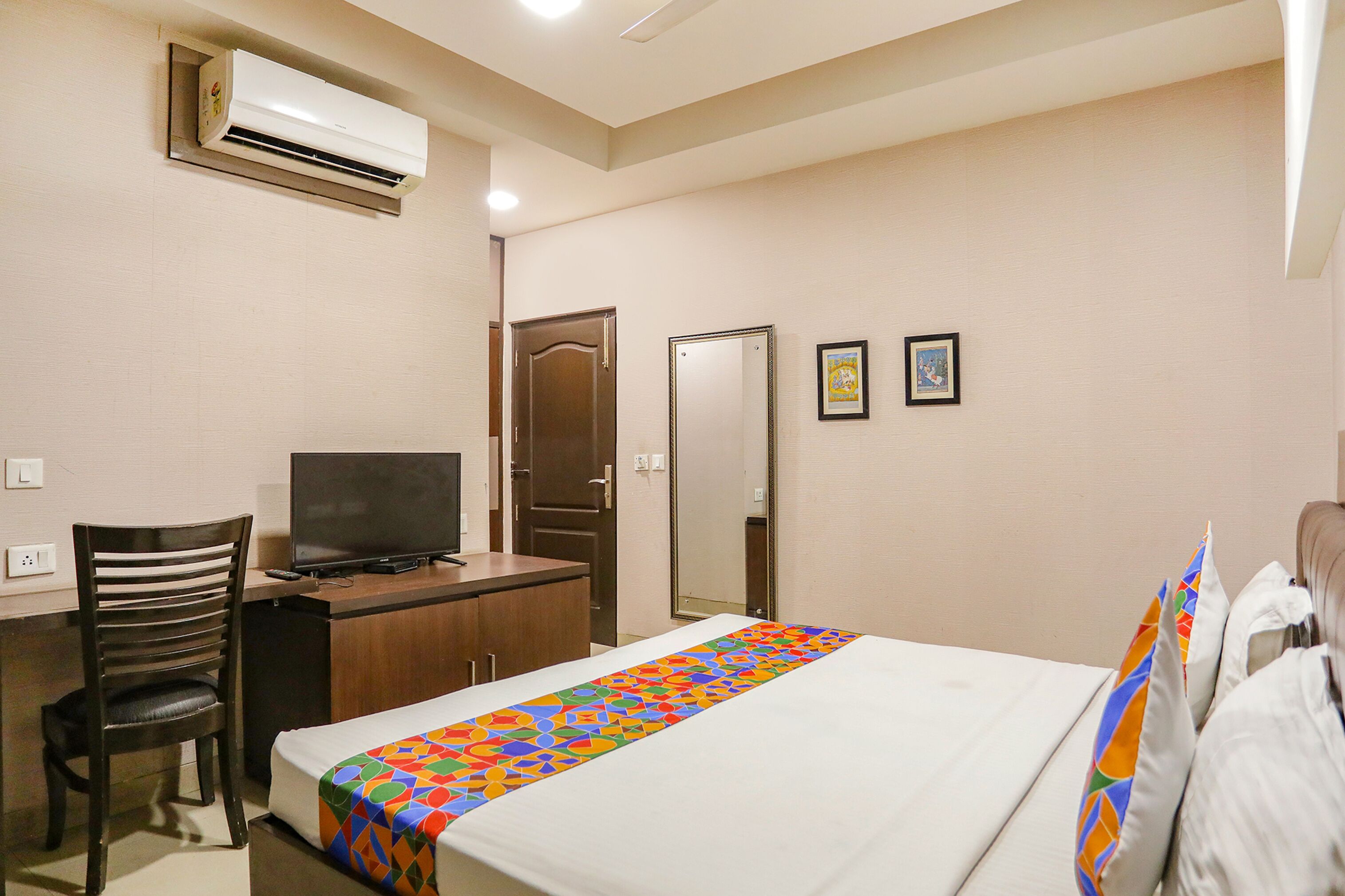A hotel room with a bed, a desk with a television, a chair, a wardrobe, and an air conditioning unit.