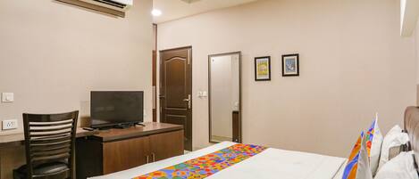 A hotel room with a bed, a desk with a television, a chair, a wardrobe, and an air conditioning unit.