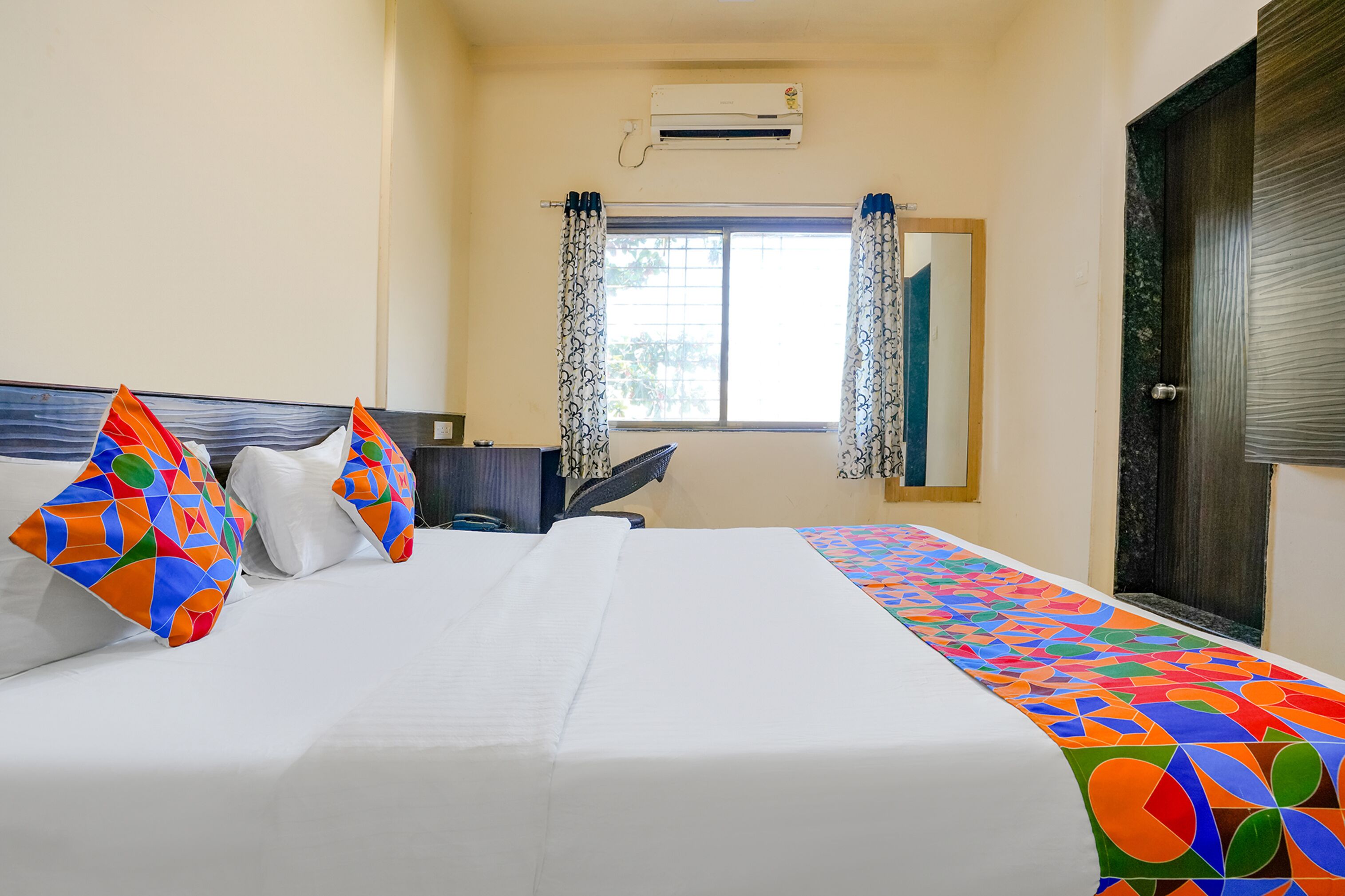 A hotel room with a bed, pillows, a window with curtains, an air conditioning unit, and a door.