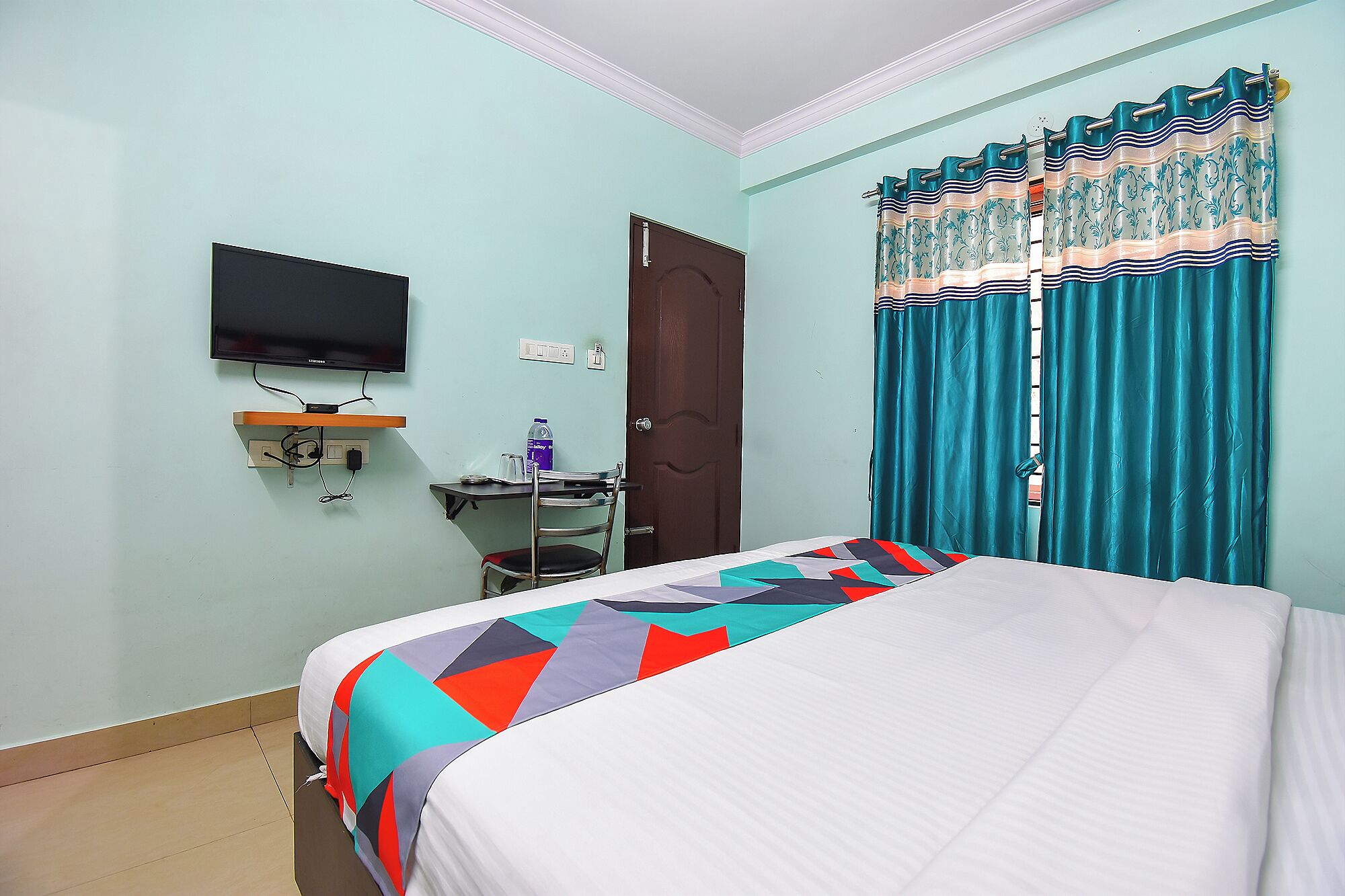 Quarto com cama, televisão na parede, escrivaninha com cadeira e porta.