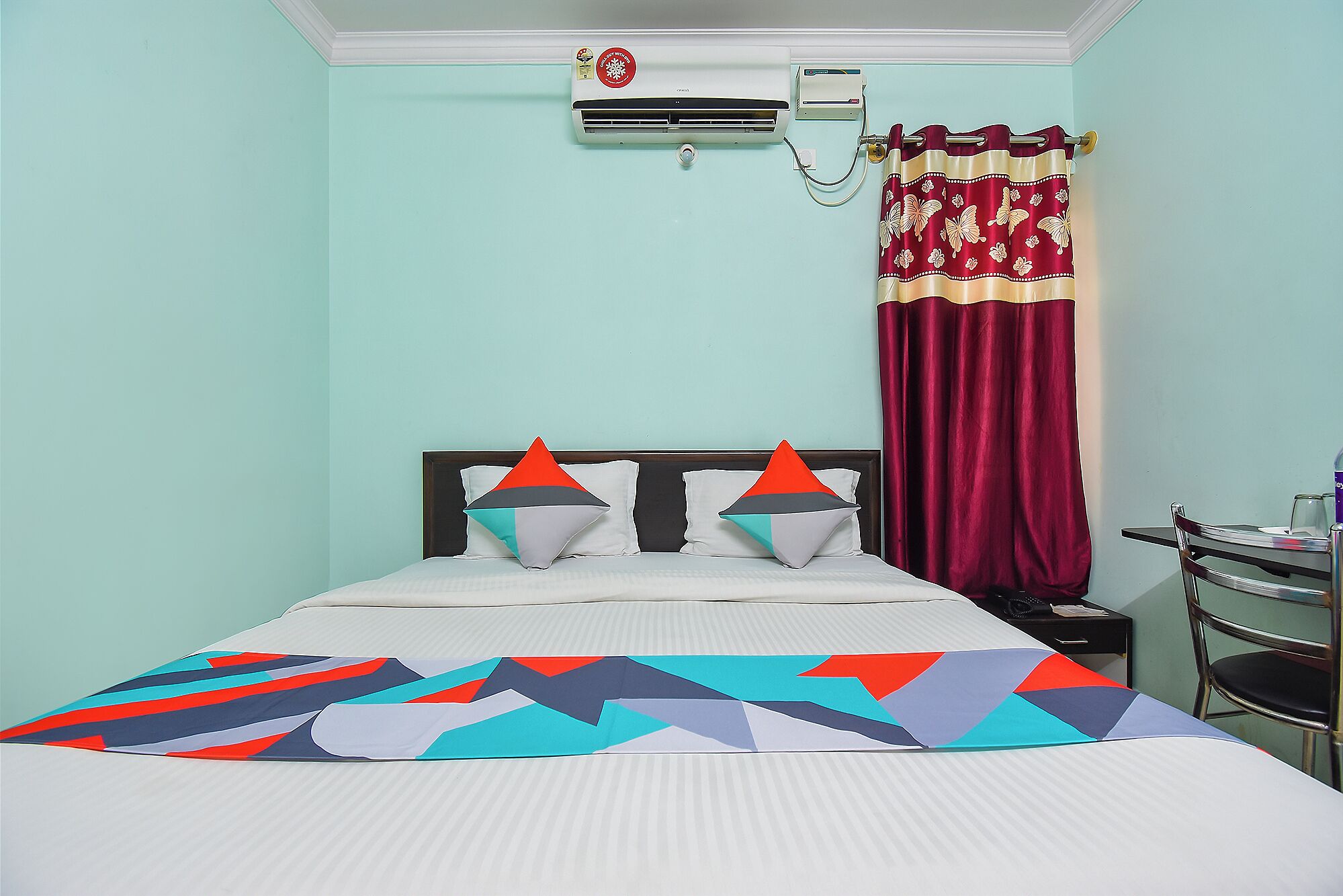 Habitación de hotel con cama, almohadas, una colcha, unidad de aire acondicionado, cortina y escritorio con silla.