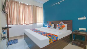 A bedroom with a bed, a bedside table, a nightstand, a window with curtains, and a wall-mounted light fixture. - FabHotel Saket Inn (Bhopal)