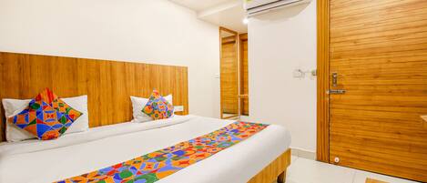 Une chambre d’hôtel avec une tête de lit en bois, un grand lit recouvert d’une courtepointe aux motifs géométriques colorés, un mur blanc et une porte en bois.