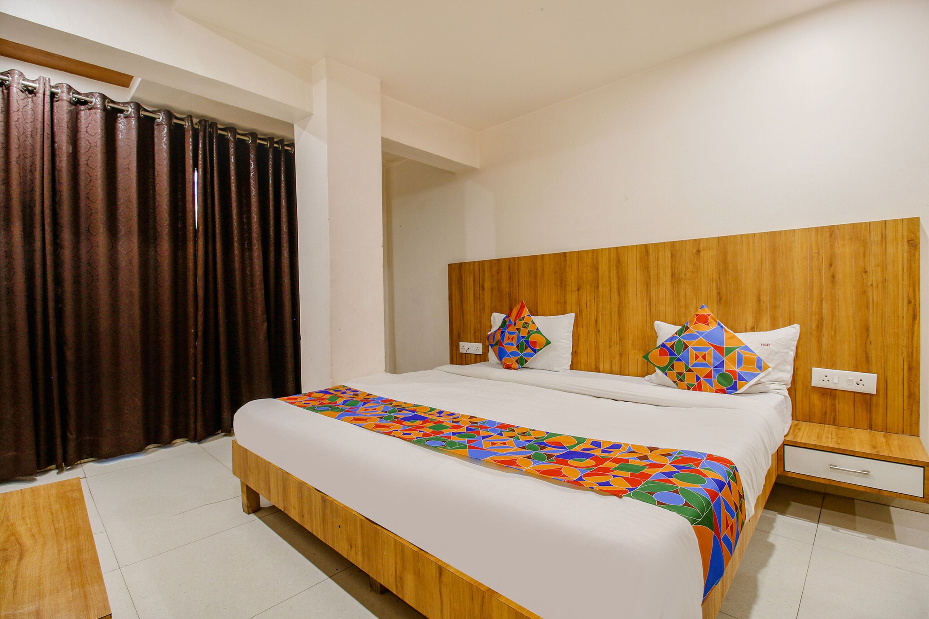 A hotel room with a wooden bed frame, a white mattress, a colorful geometric-patterned bedspread, a wooden nightstand with a drawer, and a brown curtain.