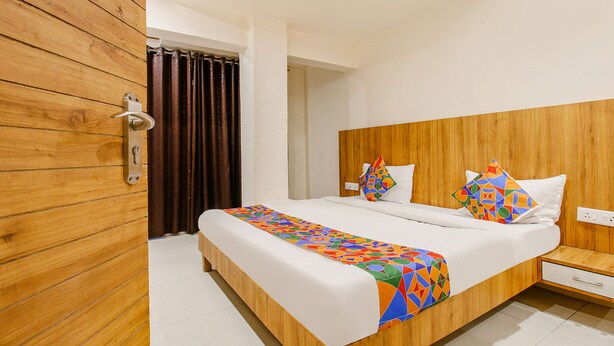 A hotel room with a wooden headboard, a large bed with a colorful geometric-patterned bedspread, a wooden nightstand, and a white wall. - Fabexpress Sunshine Regency (Bhopal)