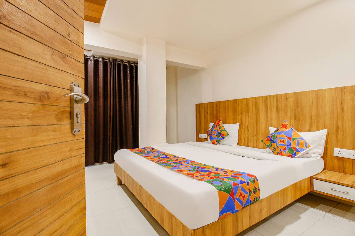 A hotel room with a wooden headboard, a large bed with a colorful geometric-patterned bedspread, a wooden nightstand, and a white wall.