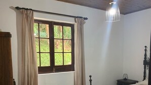 Een slaapkamer met een houten plafond, een raam met gordijnen, een bed, een nachtkastje en een stoel.