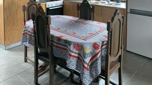 Een keuken met houten kastjes, een eettafel met een tafelkleed met dessin, en een raam met uitzicht op groen.