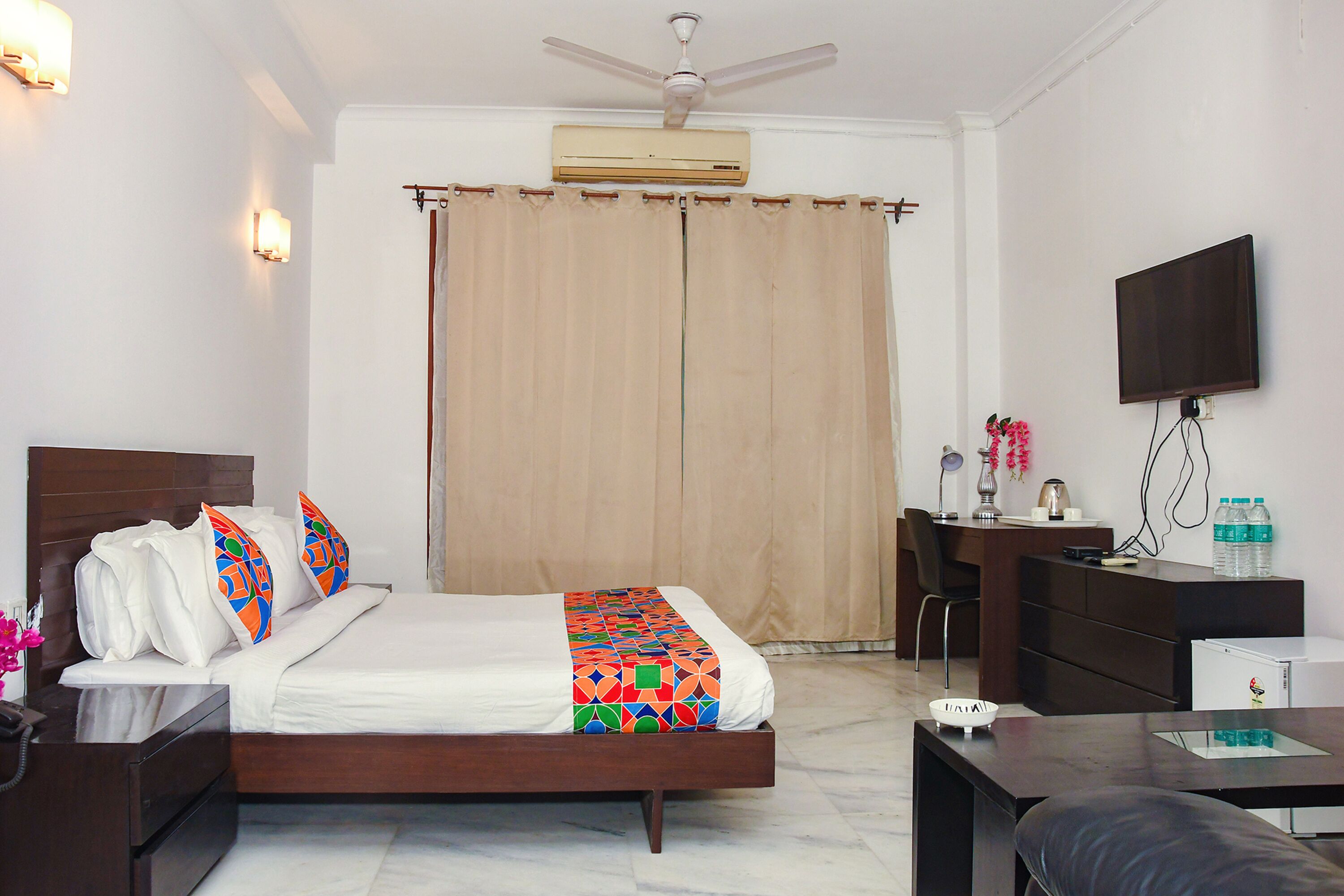 A hotel room with a bed, bedside table, desk, chair, TV, and an air conditioning unit.