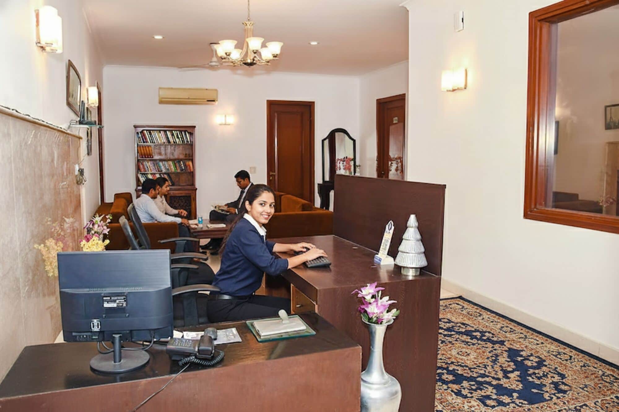 A reception desk with a computer, a phone, and a vase with flowers. There is a bookshelf with books and a chandelier above.