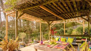 A covered outdoor seating area with a wooden pergola, a table with colorful placemats, and potted plants. - Dans la forêt de Pin by Interhome (Arcachon)
