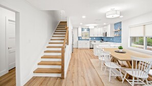 A modern kitchen with white cabinets, a wooden staircase, and a dining area with a wooden table and white chairs.