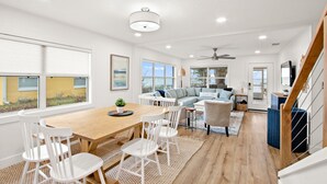 A spacious living area with a wooden dining table, white chairs, a grey sofa, and a ceiling fan.