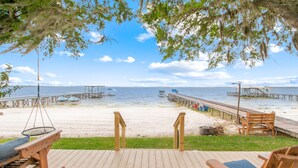 A wooden deck with two rocking chairs and a swing, overlooking a beach with a pier and boats.