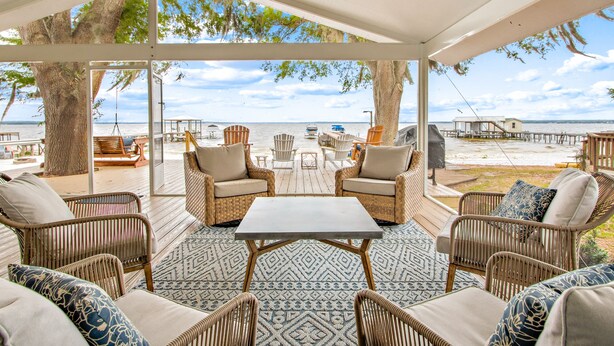 A covered outdoor seating area with wicker furniture, a table, and a view of the beach and water.