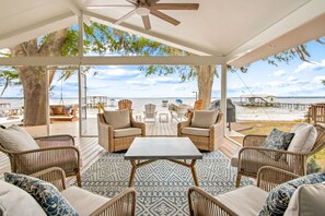 A covered outdoor seating area with wicker furniture, a table, and a view of the beach and water.