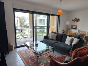A modern living room with a glass coffee table, a sofa with patterned cushions, and a view of a courtyard with a dining area. - Newquay by Interhome (Dinard)