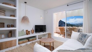 Ein modernes Wohnzimmer mit Fernseher, Regalen mit Dekoration, einem runden Holztisch und einer großen Schiebeglas-Tür, die zu einem Sitzbereich im Freien mit Landschaftsblick führt.