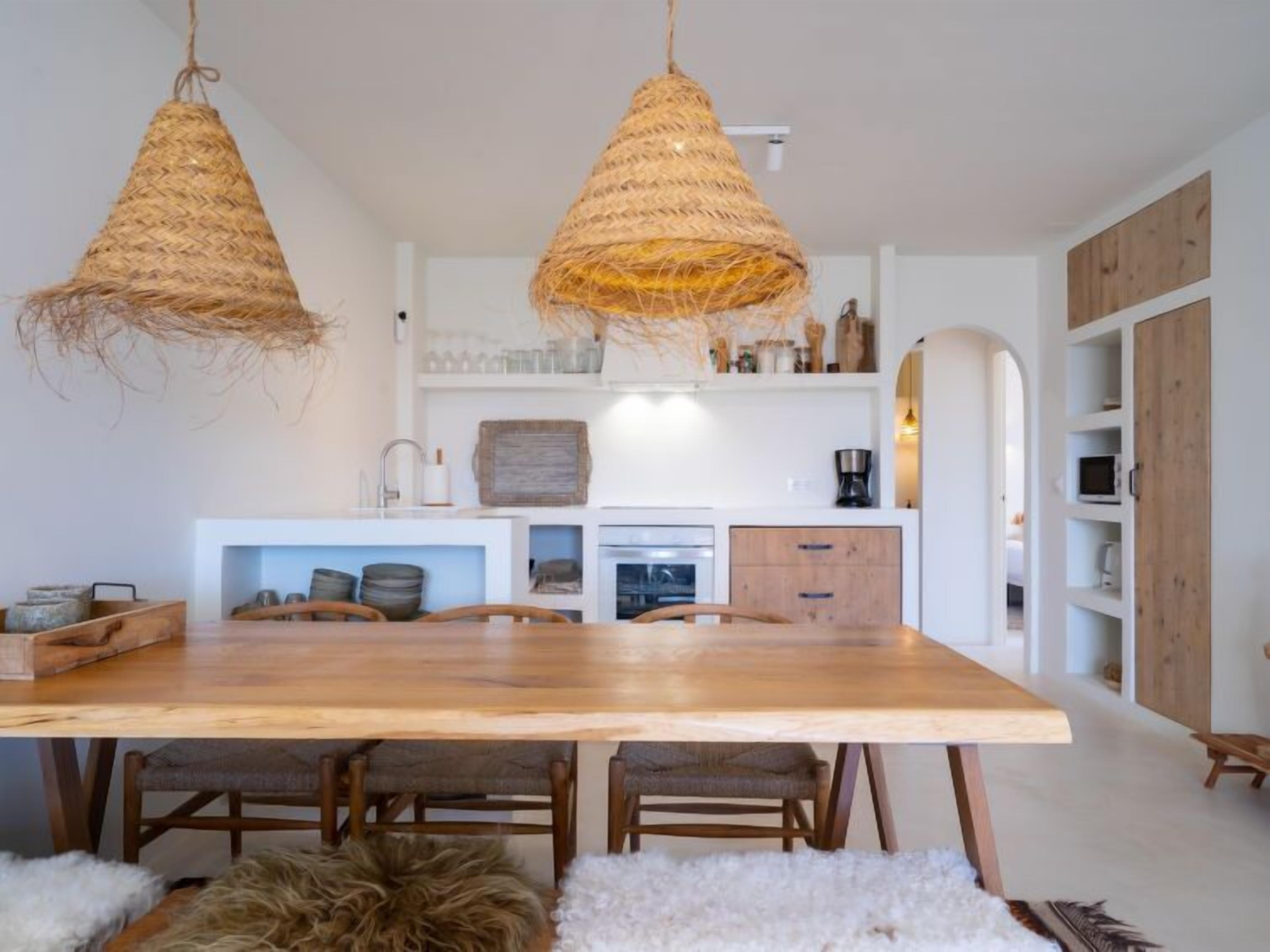 A modern kitchen with a wooden dining table, two hanging woven light fixtures, and a white interior.