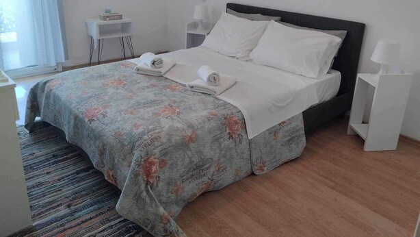 A neatly made bed with white linens and a floral-patterned duvet, a bedside table with a lamp, and a small shelf with a book. - Apartment - Sleeps 4 & Free Parking (Chania)