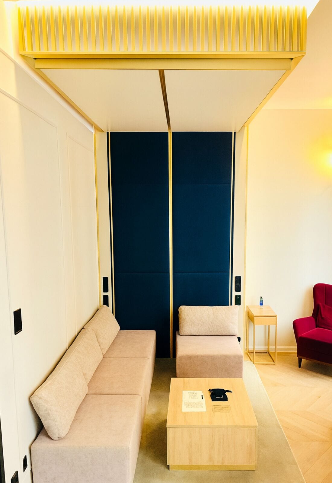 A modern hotel room with a sofa, a smaller seating area, a wooden coffee table, and a red chair.