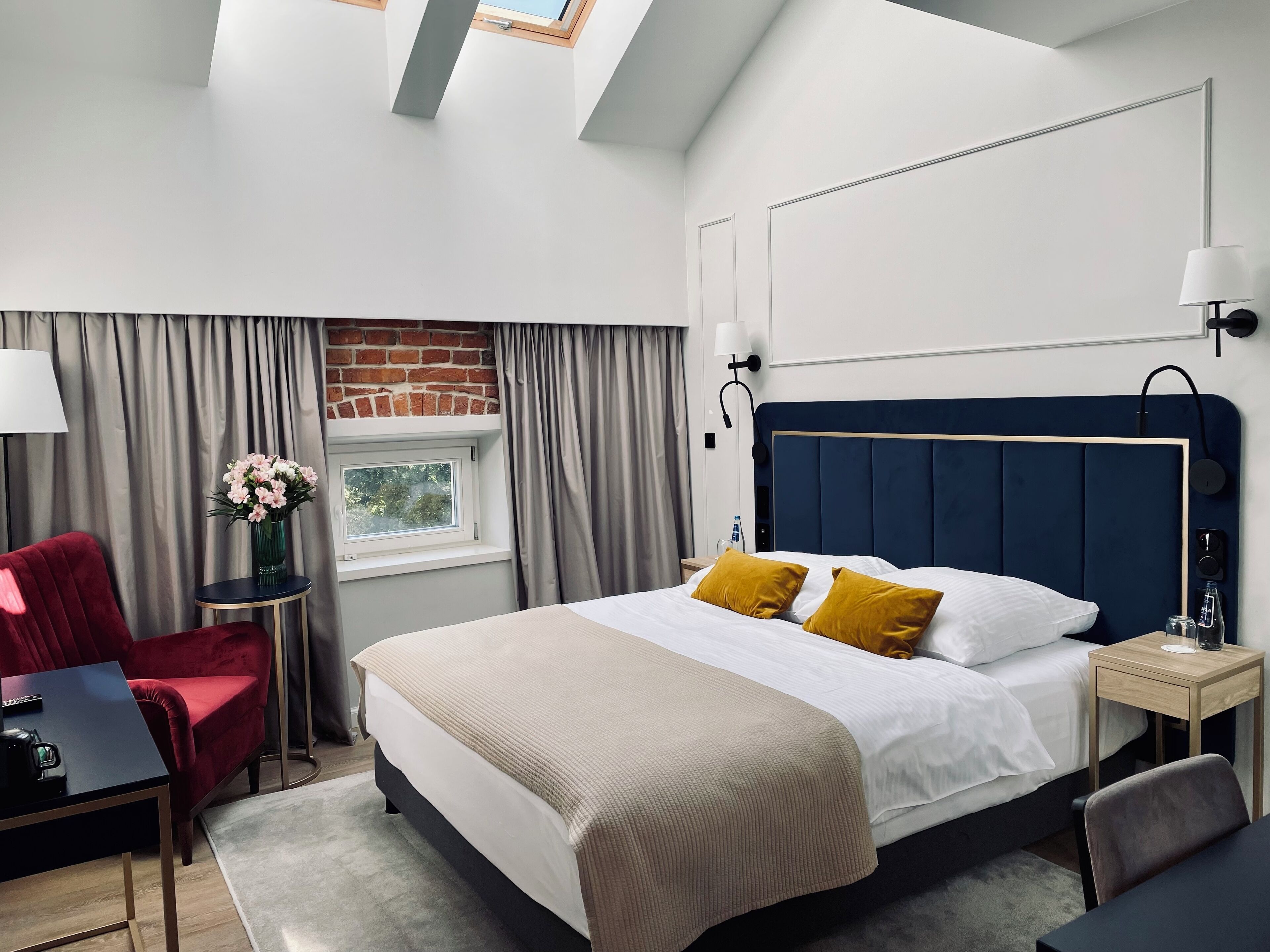 Quarto de hotel moderno com uma cama grande, uma escrivaninha, uma cadeira, uma mesinha de centro com um vaso de flores e uma janela com cortinas.