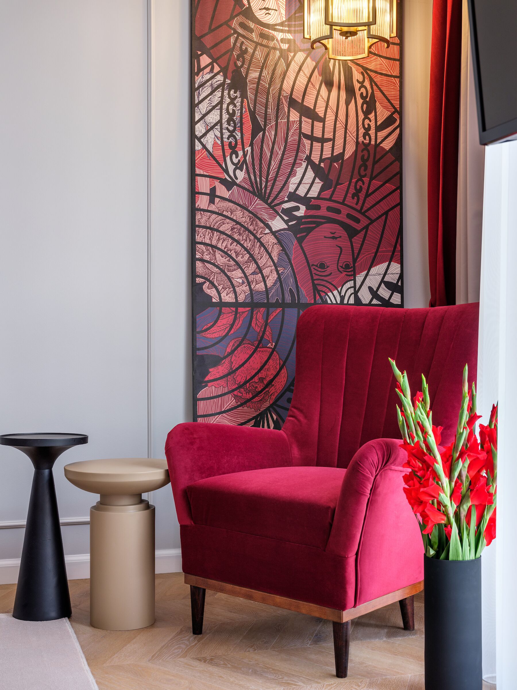 Un fauteuil rouge, une table ronde et un grand vase orné de fleurs rouges, dans une pièce dont le mur est décoré d’un motif et qui est éclairée par un luminaire.