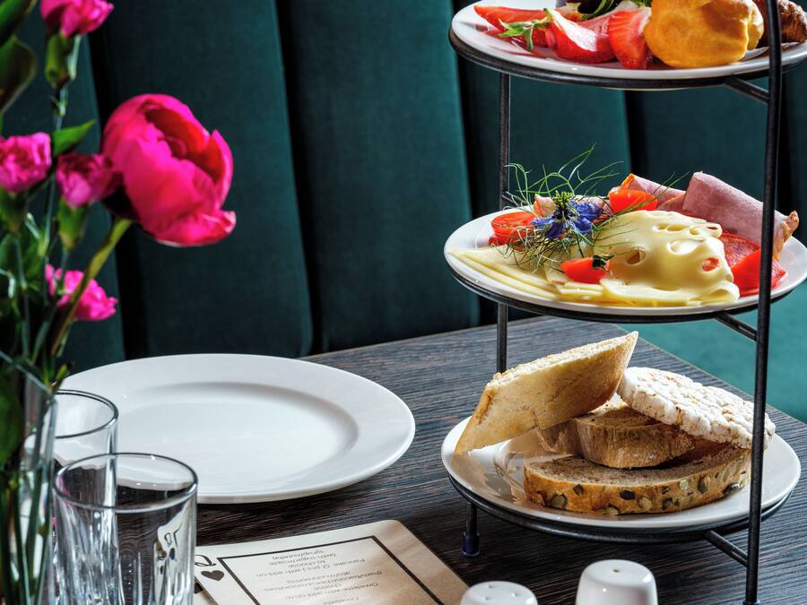 Un expositor de tres niveles con diversos pasteles y frutas, un florero con flores rosas, y un menú sobre una mesa con platos y vasos.
