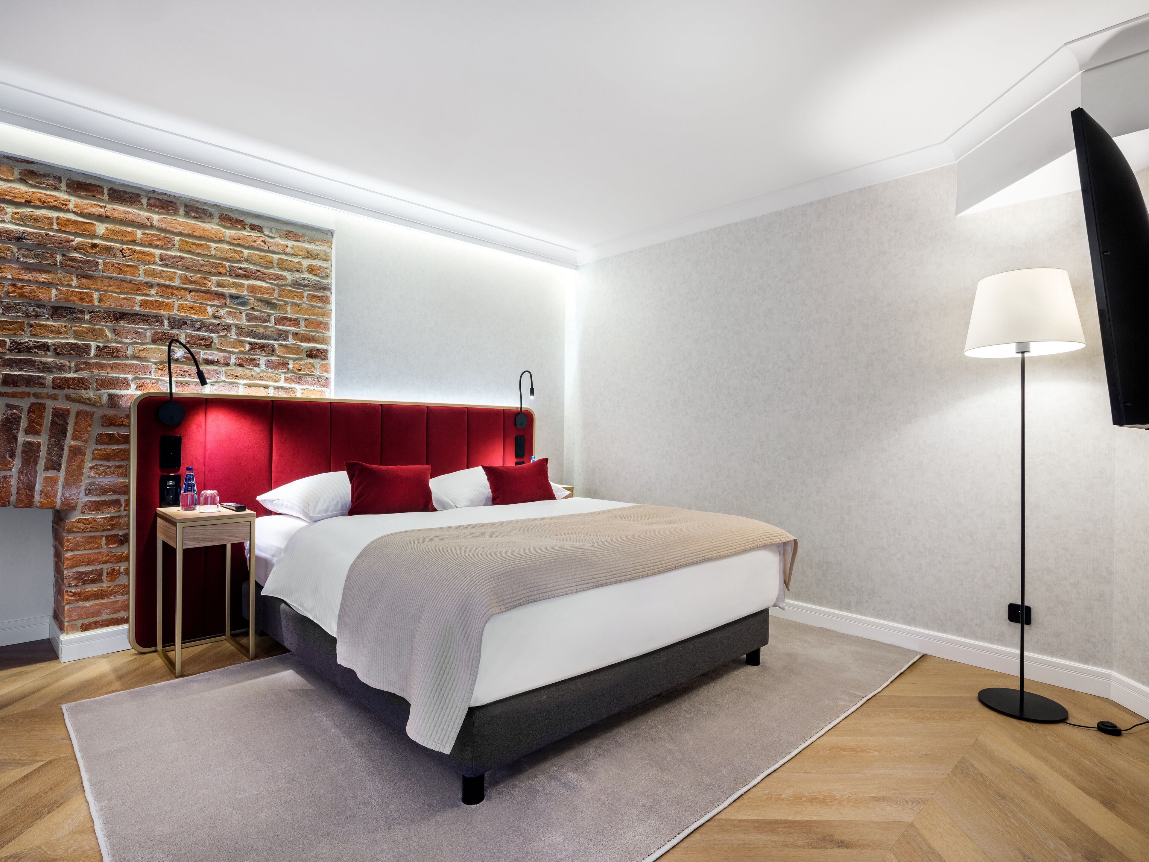A modern bedroom with a brick accent wall, a red upholstered headboard, a bedside table with a lamp, and a floor lamp.