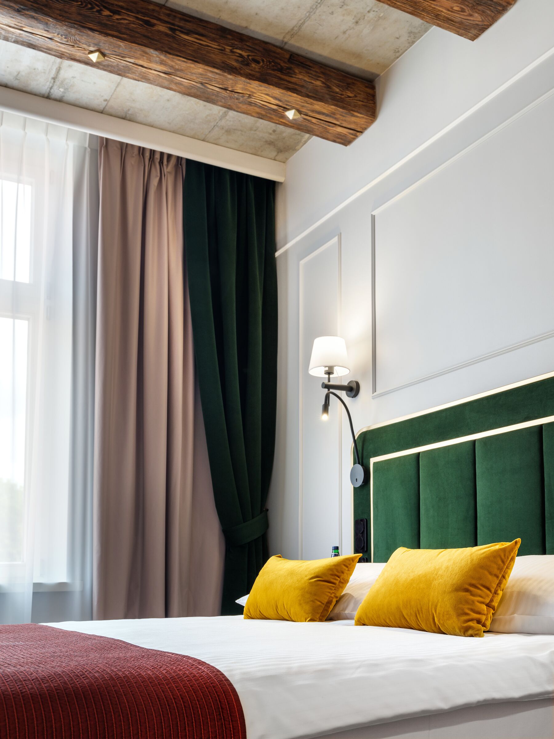 Habitación de hotel con cama, cabecera tapizada de verde, almohadas amarillas, lámpara de noche y ventana con cortinas.