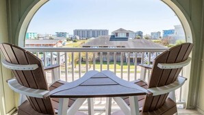 Uma varanda com duas cadeiras Adirondack e uma mesa pequena, com vista para um horizonte urbano com edifícios e uma área gramada.
